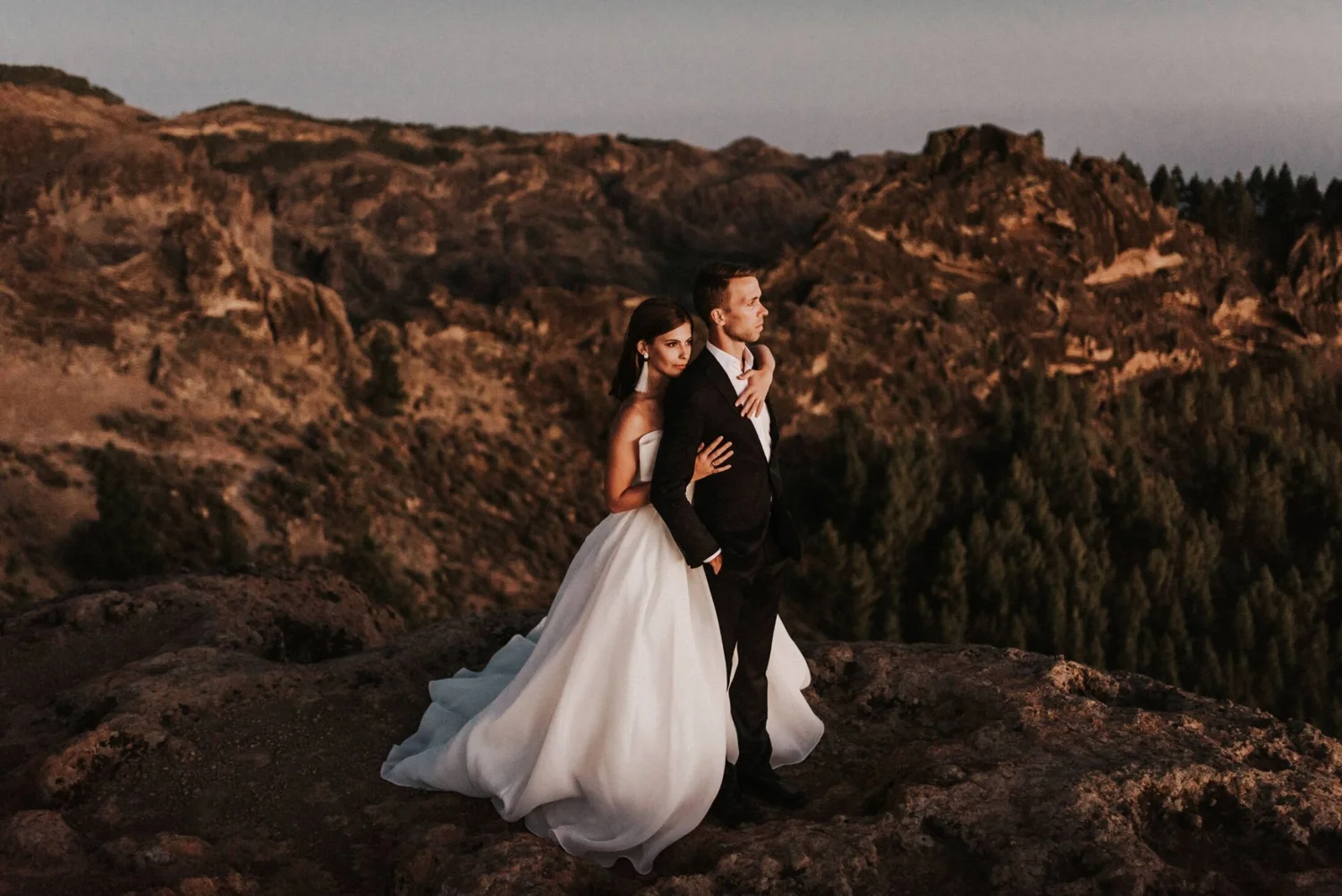 A bride and groom standing embraces on rocky terrain with rugged mountains in the background during twilight.