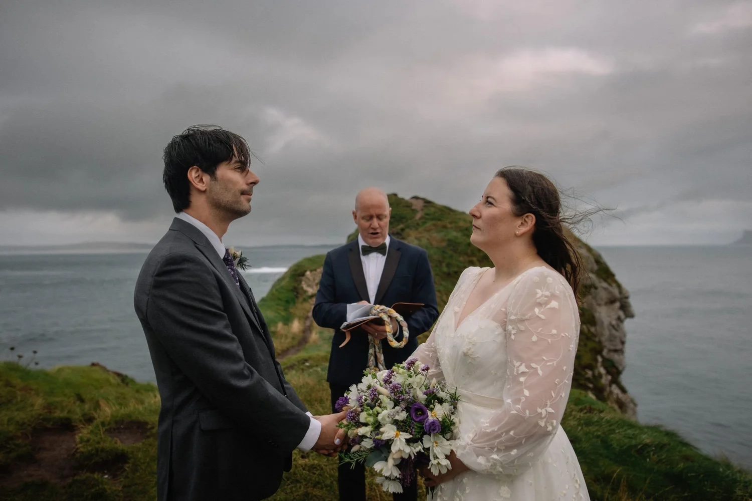 A wedding ceremony outdoors by the ocean on a cloudy day. A groom in a gray suit and a bride in a white lace dress face each other holding hands with a bouquet of white and purple flowers. The officiant, in a dark suit with a bow tie, stands behind t