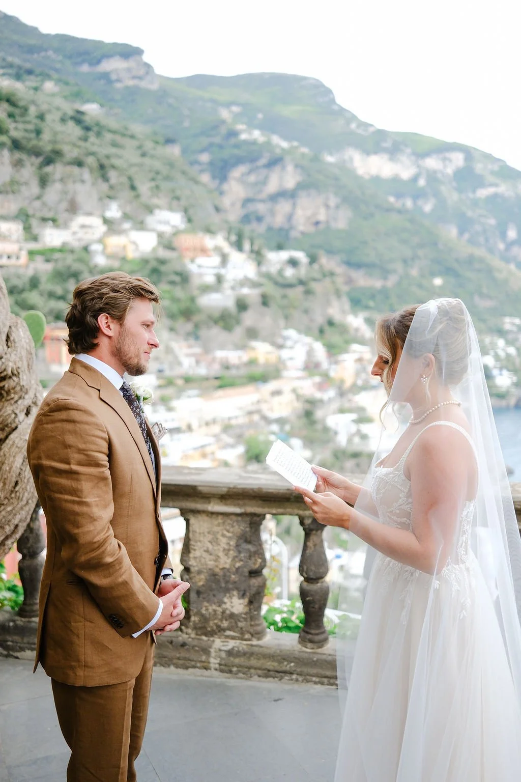 A couple getting married outdoors with a scenic hillside village in the background. The bride is reading vows, wearing a white wedding dress and veil, while the groom stands in a brown suit.