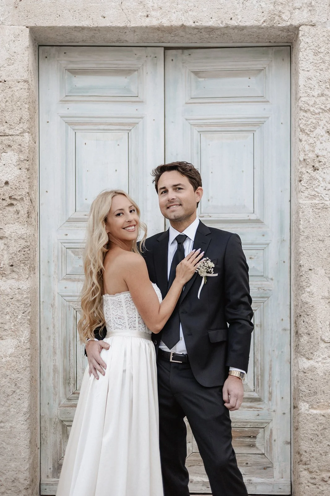 A bride and groom standing in front of a rustic wooden door, smiling and embracing each other.