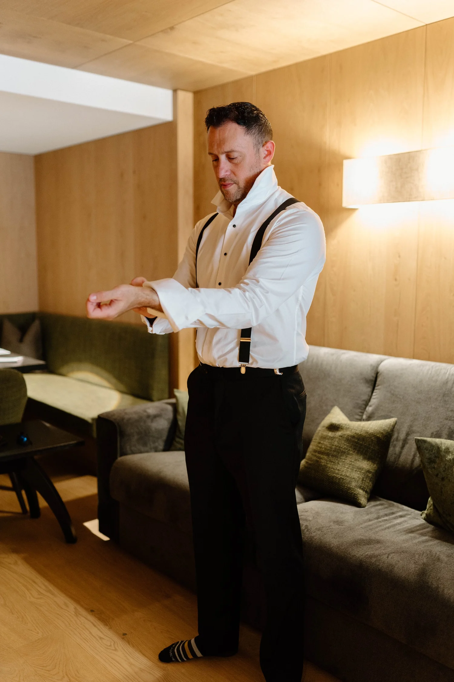 Man in white dress shirt adjusting his sleeve in a living room with wooden walls, a grey couch, and decorative pillows.