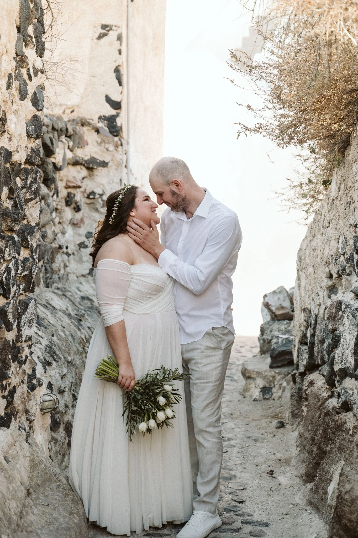 A bride and groom are embracing in a narrow stone passageway with rocky walls, overlooking the ocean.