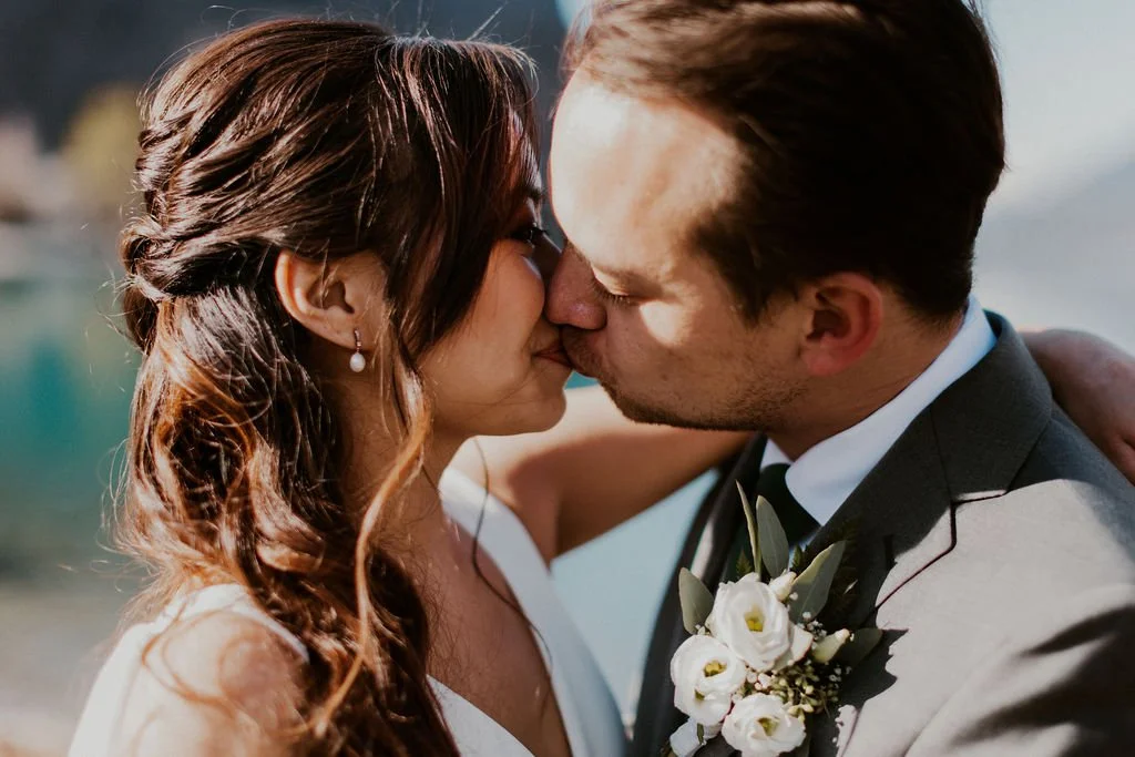 A bride and groom sharing a kiss outdoors during their wedding, with the bride wearing pearl earrings and the groom in a suit with a boutonniere.