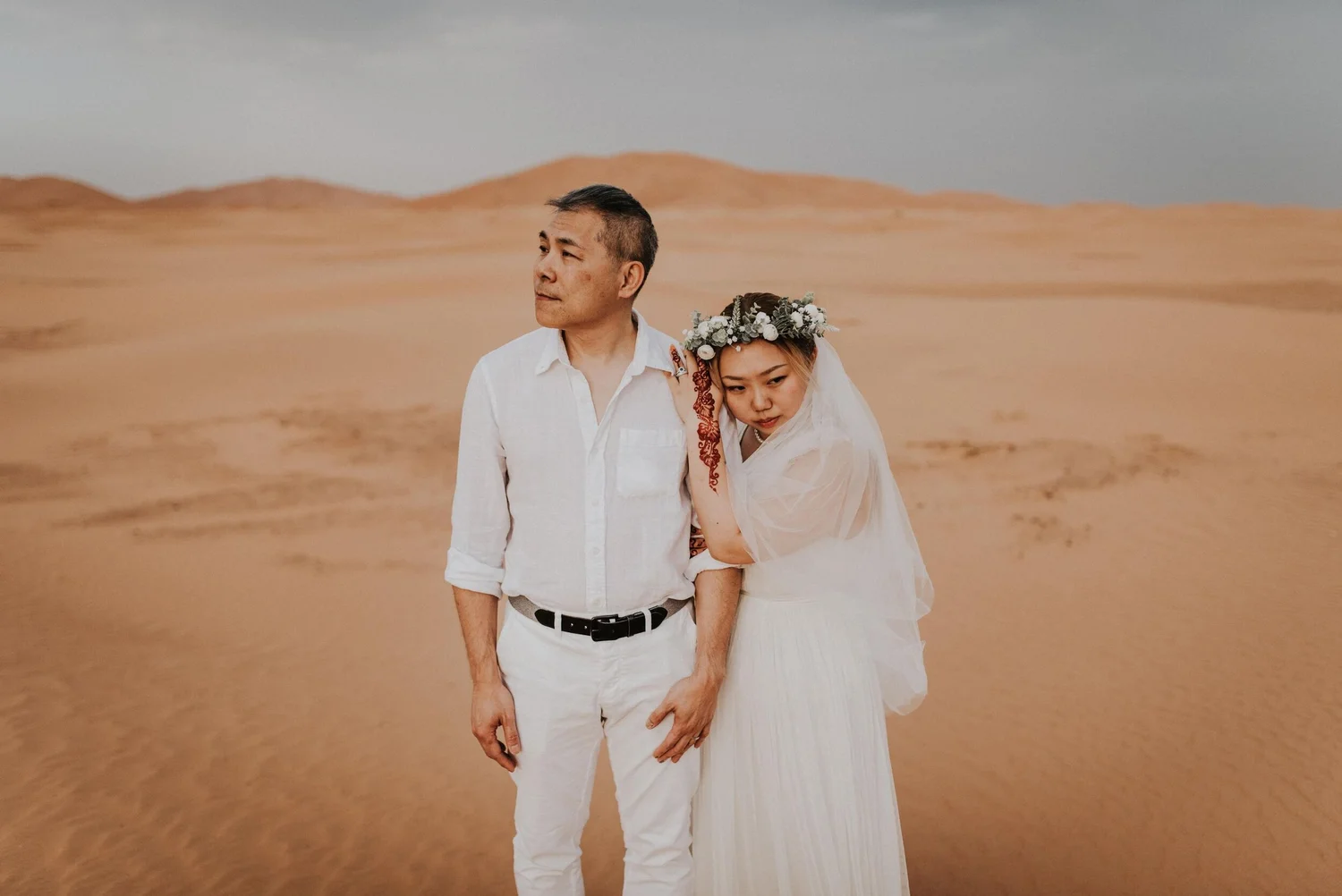 A man and woman are standing together in a desert landscape, both wearing white clothing. The woman is resting her head on the man’s shoulder, and she has a floral crown on her head.