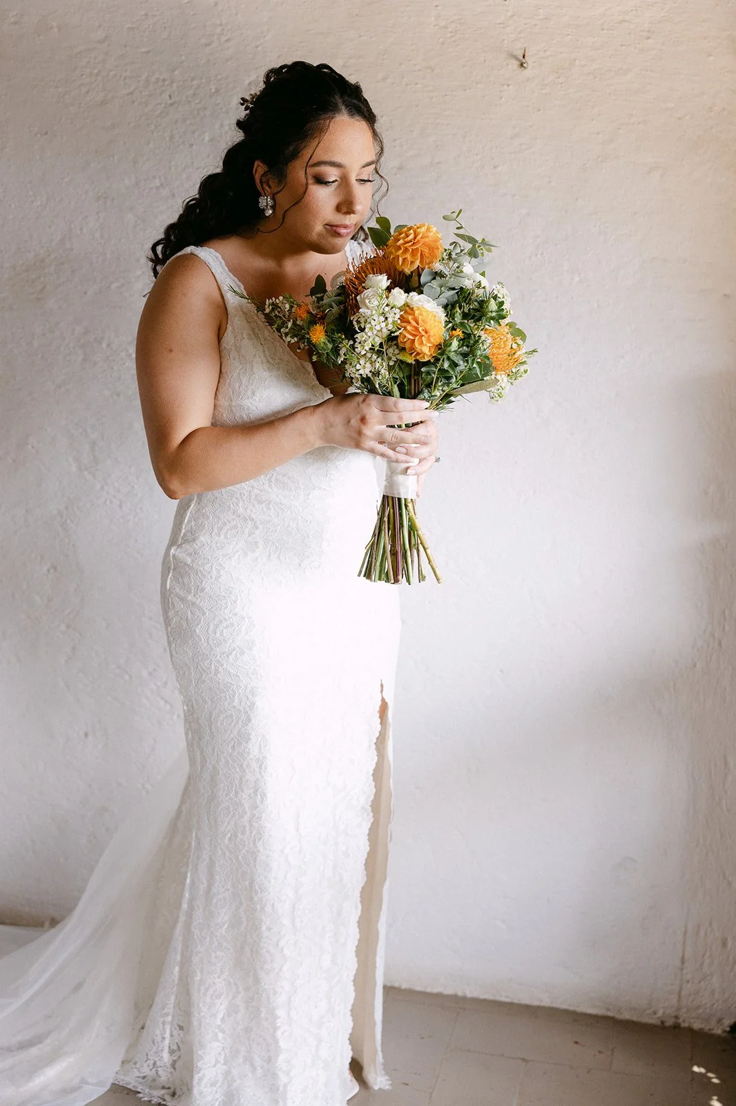 A bride in a white lace gown holding a bouquet of orange, white, and yellow flowers standing against a plain wall.