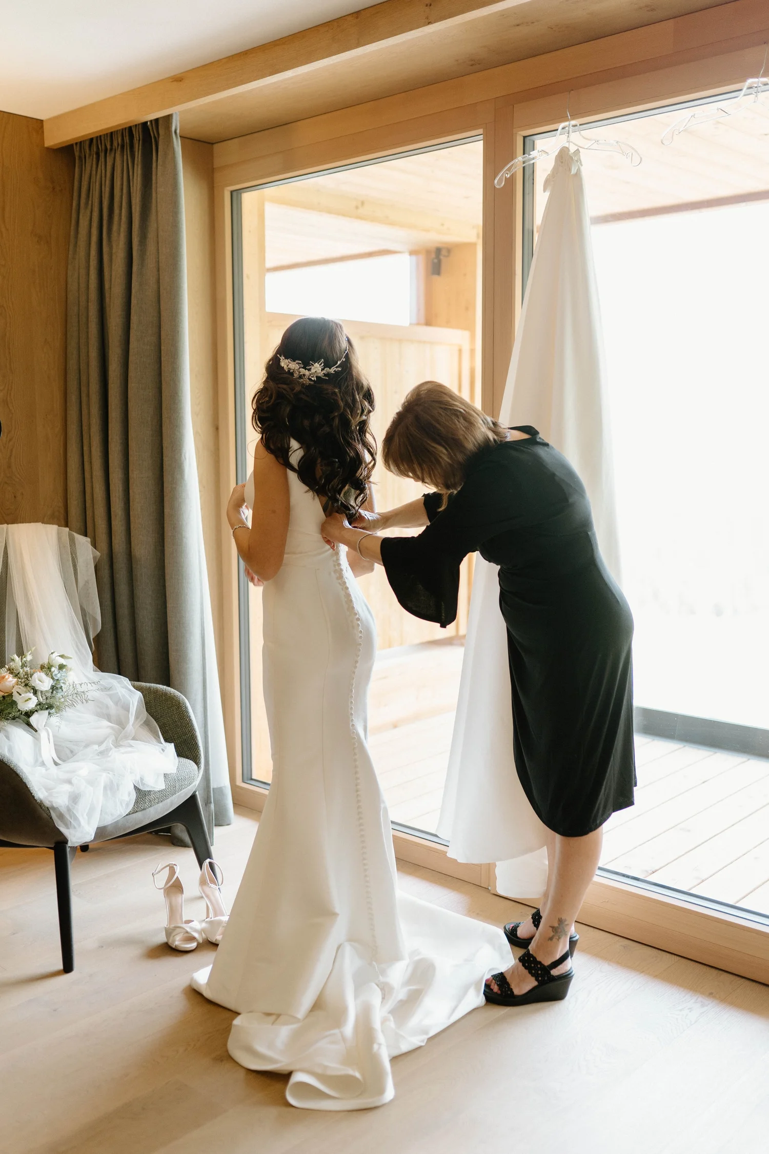 A bride with long dark curly hair and a floral hairpiece is getting ready in a wooden room with large windows. Someone is helping her with her wedding dress. Shoes and a bouquet are on the floor and nearby.