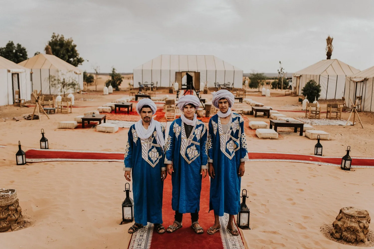 Three men dressed in traditional Moroccan kaftans with turbans, standing in a desert setting with tents and decorative lanterns.