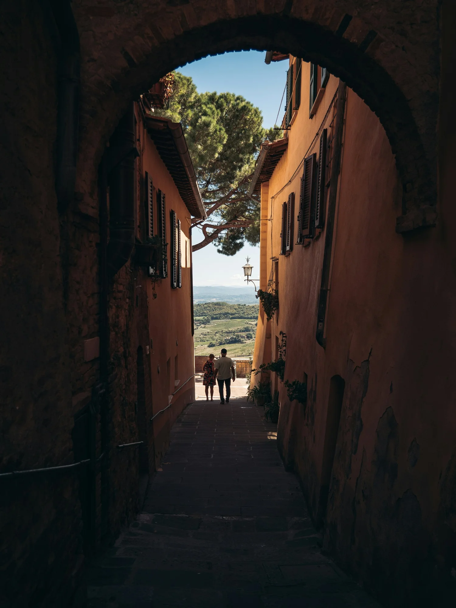 View through a dark stone archway looking out onto a narrow street with beige buildings, black shutters, a tree, and a distant landscape with hills under a clear blue sky. Two people walk away from the camera.