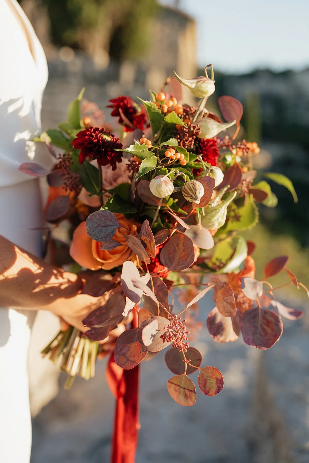 A bouquet of flowers with red dahlias, orange roses, and various greenery and foliage, held by a person wearing a white outfit outdoors.