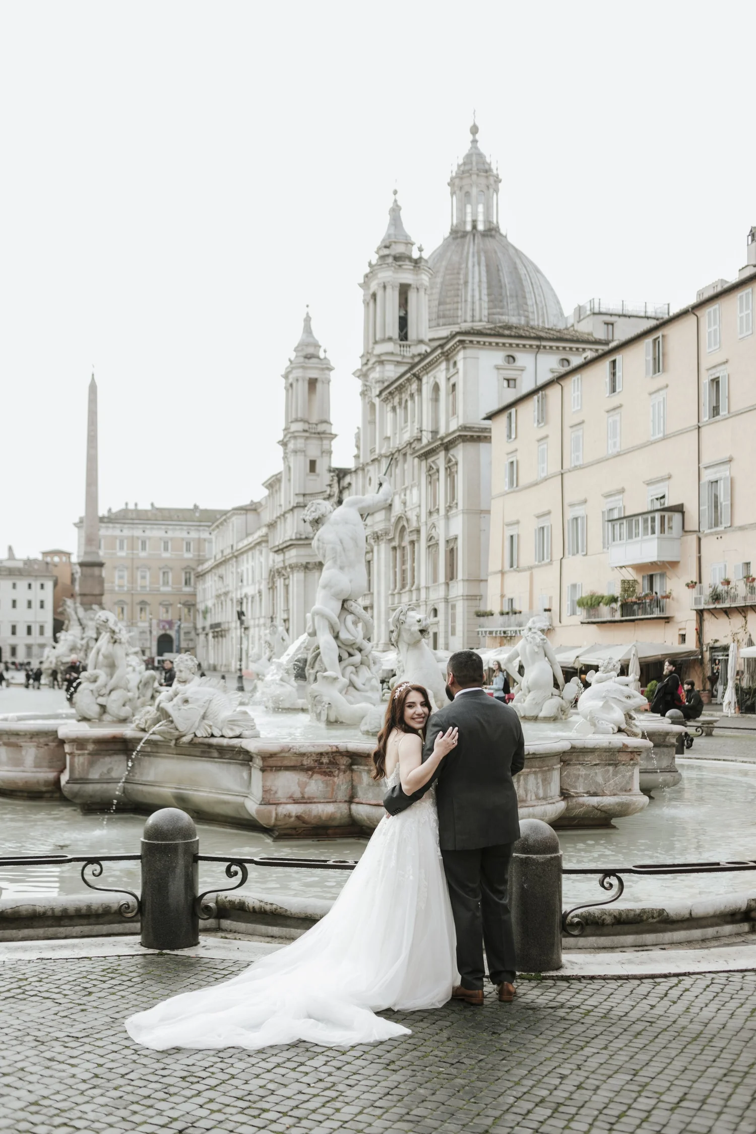 A bride and groom standing in front of a fountain with sculptures in Piazza Navona, Rome, Italy. The bride is in a white wedding dress and the groom in a dark suit, embracing each other. In the background, historic buildings and church domes are visible.