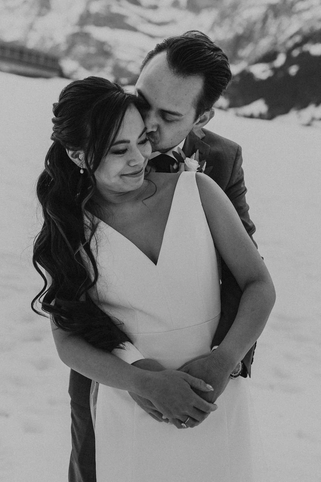 A black and white photo of a newlywed couple embracing outdoors, with the groom kissing the bride's temple, in a snowy mountain setting.