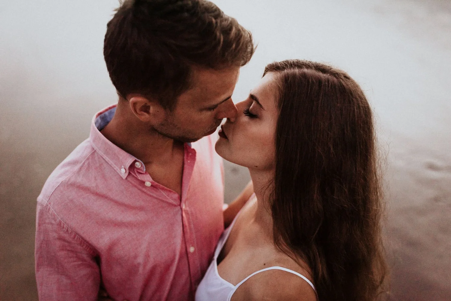 A man and woman about to kiss, standing close on a beach with a blurred background.
