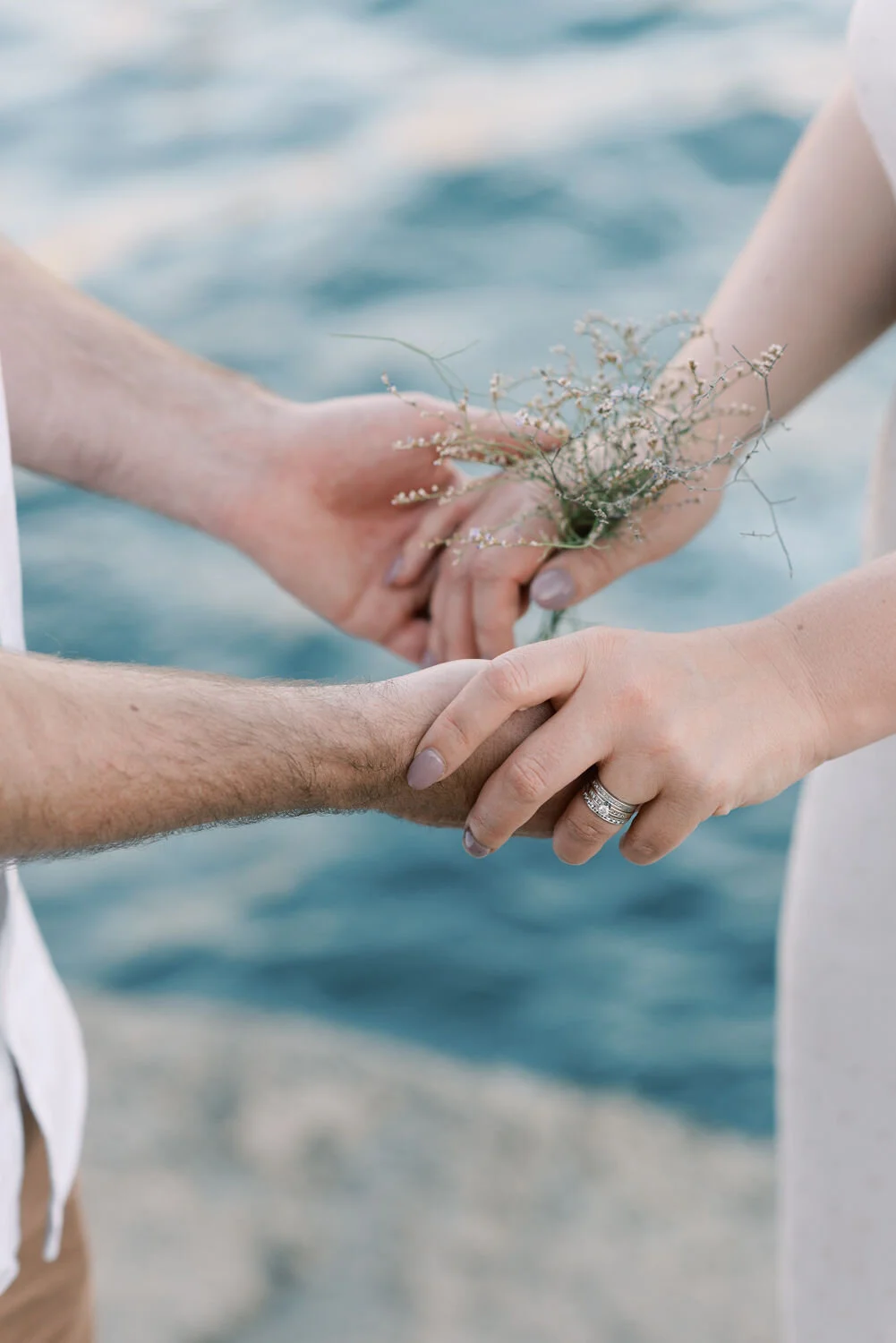 A couple holding hands, with the woman wearing an engagement or wedding ring, by the water, with one person holding a small bouquet of wildflowers.