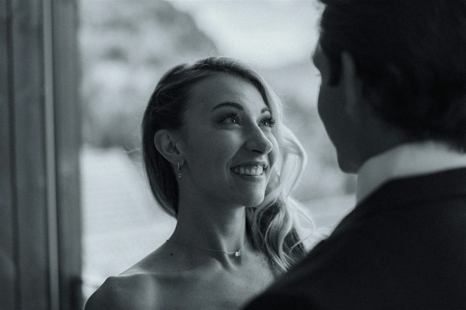 A black and white photo of a smiling woman with wavy hair looking at a man whose face is partially visible, in a close-up intimate moment.