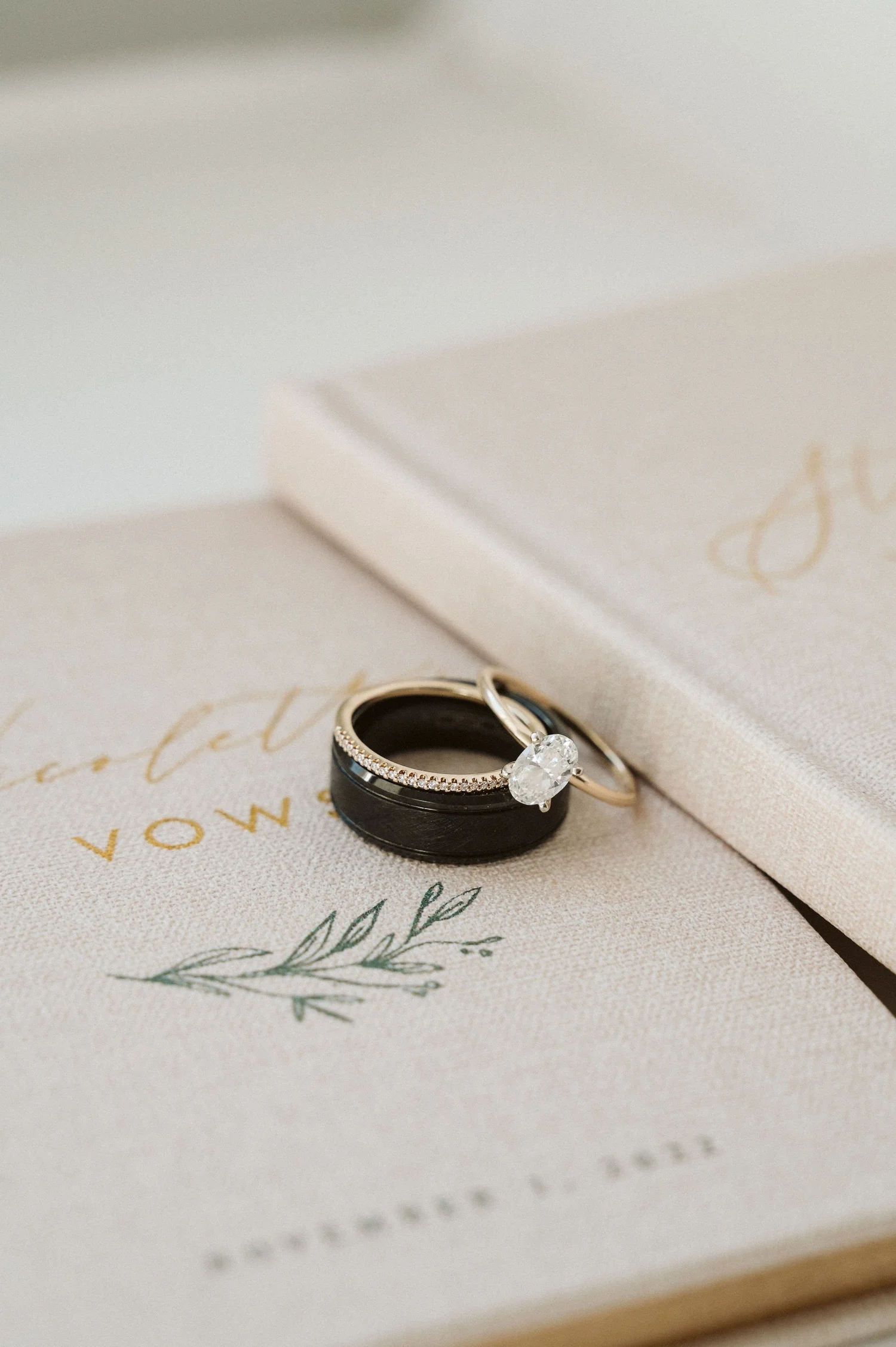 Close-up of wedding rings, including an engagement ring with a large diamond, set on top of an invitation with elegant calligraphy, on a cream-colored surface.