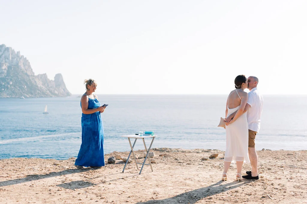A couple getting married on a beach, with a woman officiant standing nearby, overlooking the ocean and rocky cliffs in the background.