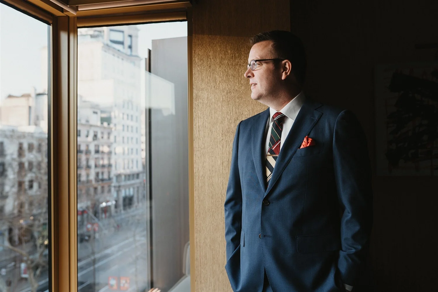 A man in a blue suit, striped tie, and glasses is standing indoors near a window, looking outside at a cityscape with buildings, trees, and streets.