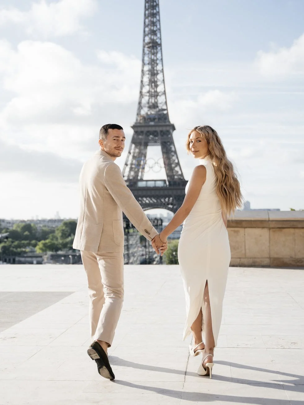 A couple holding hands and walking in front of the Eiffel Tower in Paris, France, dressed in elegant wedding attire.