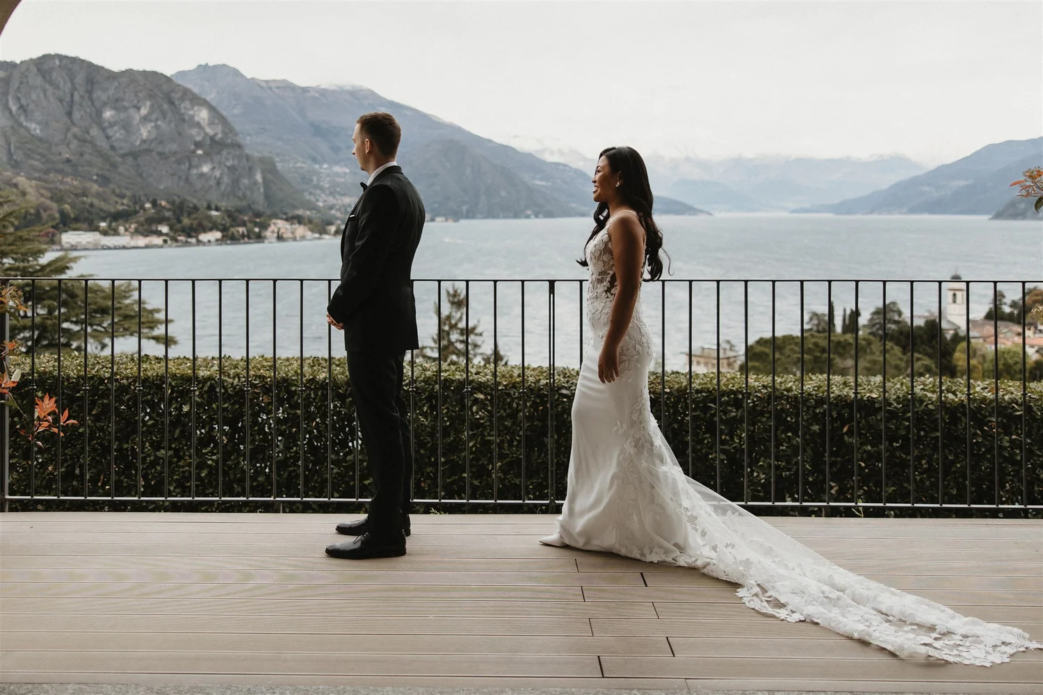 A bride and groom standing on a wooden deck near a lake with mountains in the background. The groom is dressed in a black tuxedo, and the bride is wearing a white lace wedding gown with a long train. They are facing away from each other, with the gro