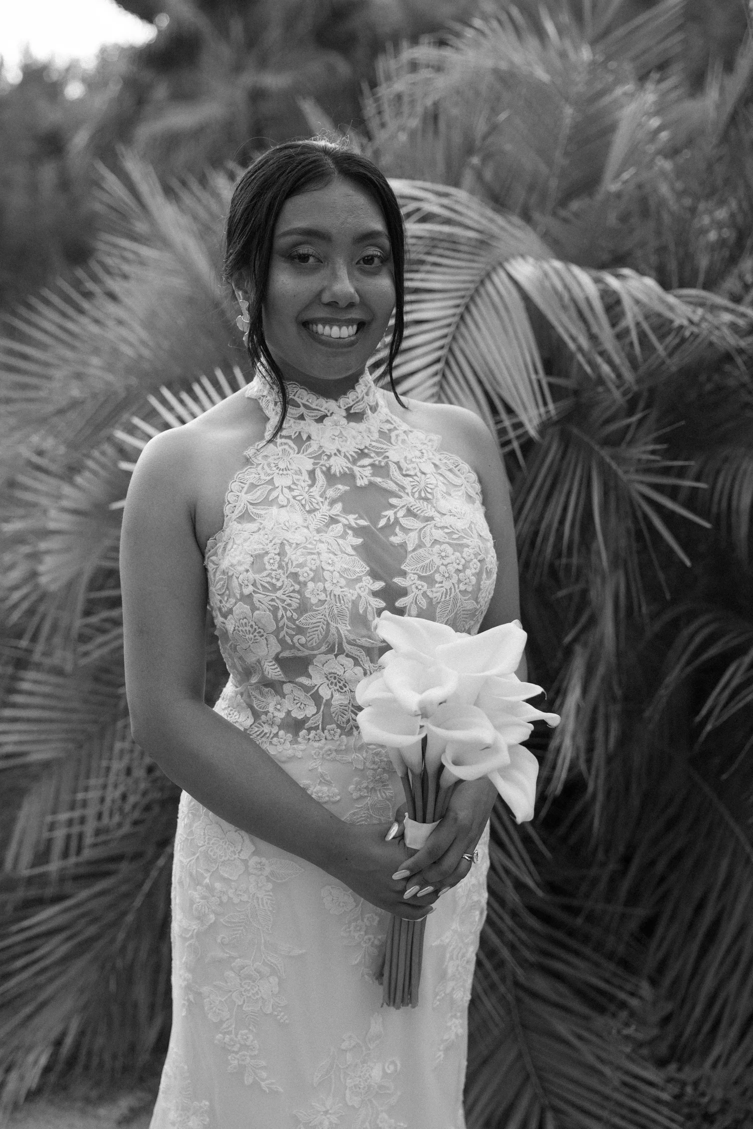 A woman dressed in a lace wedding gown, holding a bouquet of white flowers, standing outdoors surrounded by large palm leaves.
