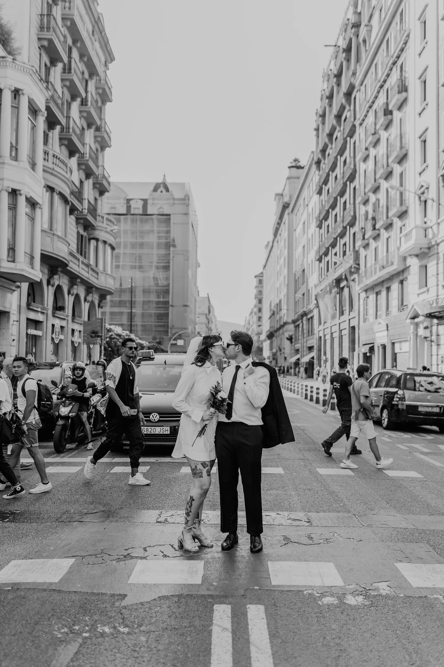 A couple dressed in wedding attire sharing a kiss in the middle of a busy city street with pedestrians and tall apartment buildings.