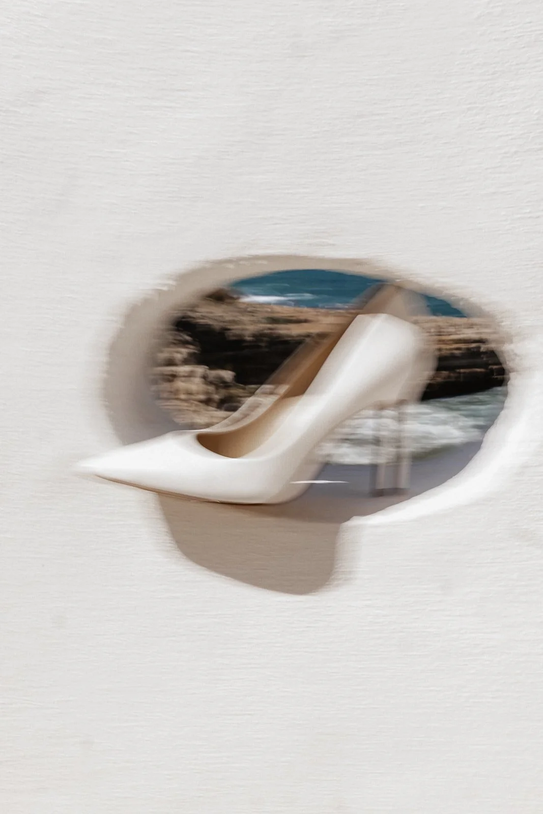 A white high-heeled shoe on a wall with a hole showing a landscape of rocks, trees, and water behind it.