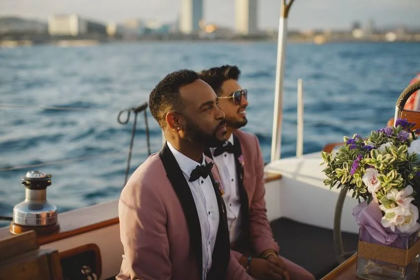 Two men dressed in pink suits and black bow ties sitting on a boat, with water and city skyline in the background.