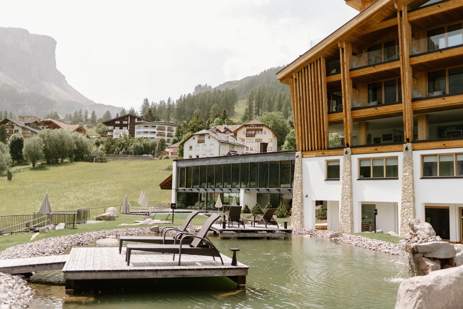 A modern multi-story building with wooden exterior and large windows, overlooking a pool area with lounge chairs and umbrellas, surrounded by lush greenery and mountains in the background.