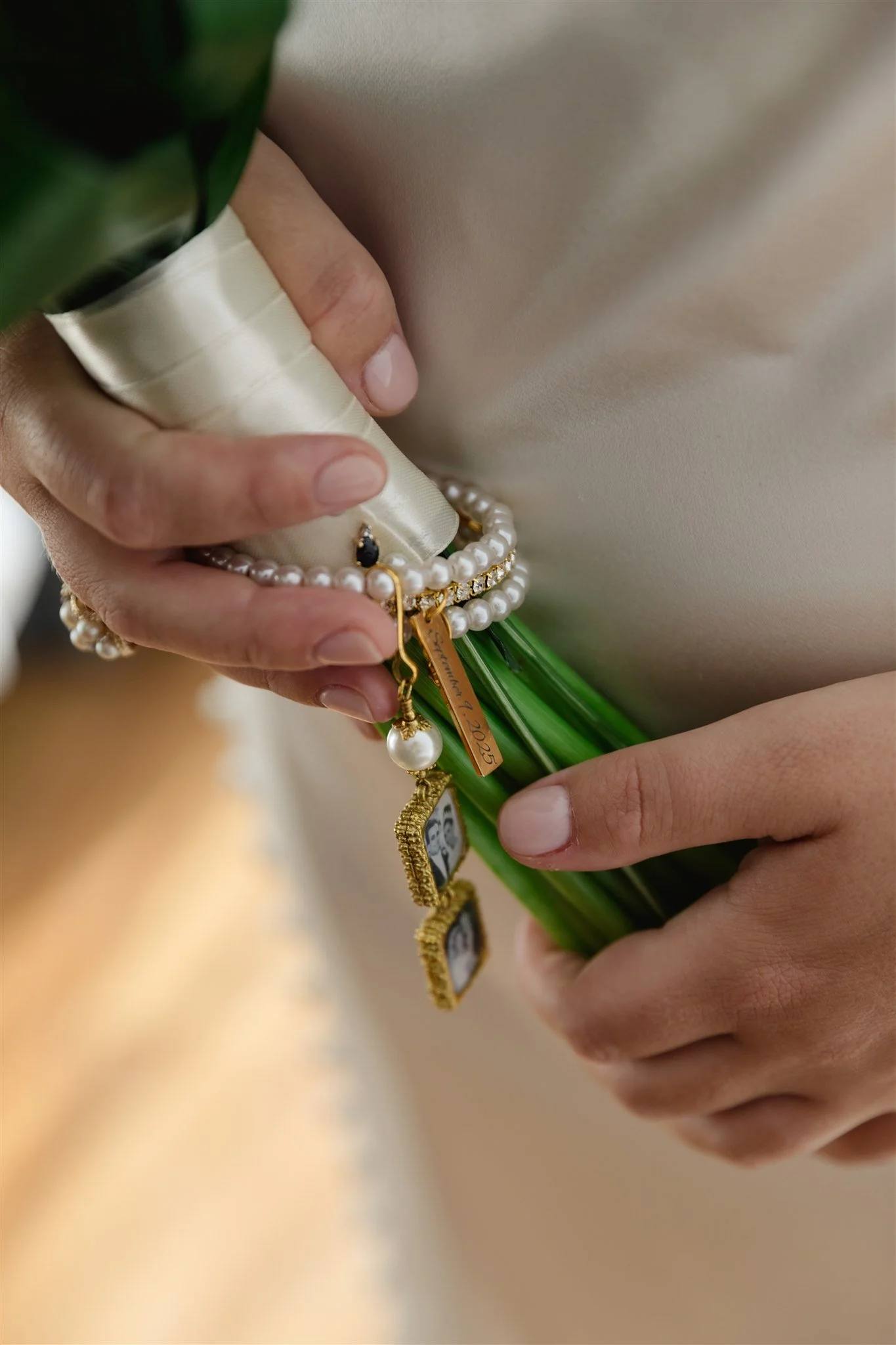 Two light-skinned hands holding a small bouquet of flowers and jewelry, including pearl necklaces, small gold charms, and a black and gold earrings. One hand has a silk white ribbon wrapped around a finger and the other hand is managing the jewelry a