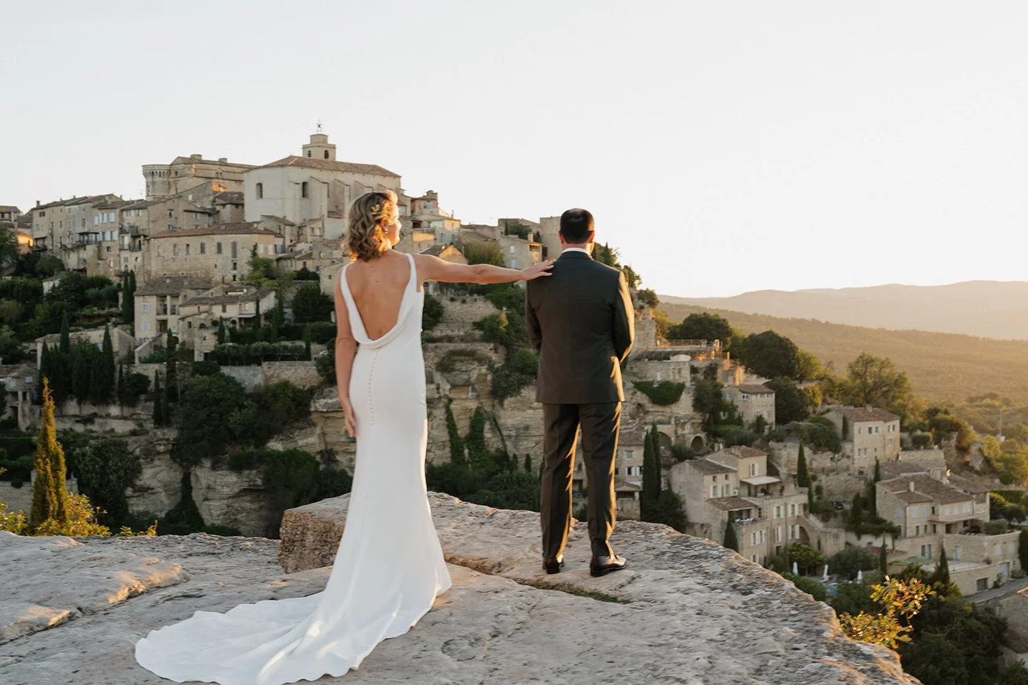 A bride in a white wedding dress with a low back and train, and a groom in a dark suit, standing on a rocky ledge overlooking a hillside town at sunset.