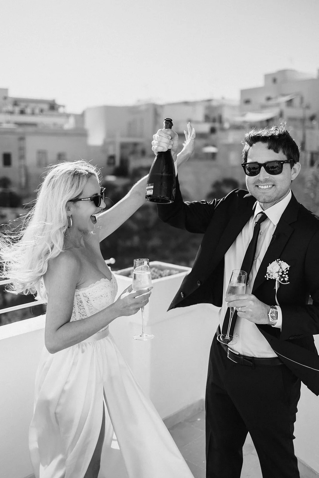 A black-and-white photo of a wedding celebration on a rooftop with a woman in a white dress and a man in a suit, both wearing sunglasses, holding champagne glasses, and clinking a bottle of champagne while smiling.