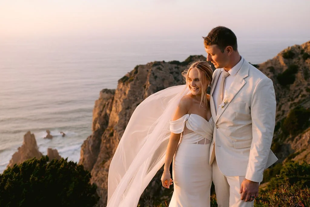 A bride and groom in wedding attire standing on a rocky cliff overlooking the ocean at sunset, smiling and looking at each other.