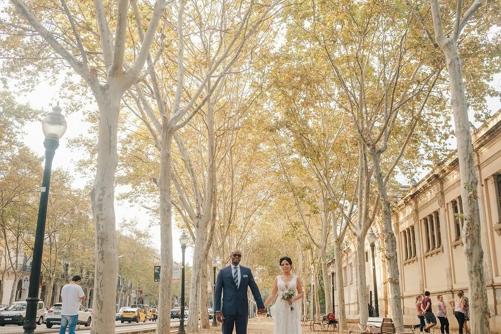 A couple in wedding attire holding hands walking down a tree-lined street with autumn foliage, classic street lamps, and historic buildings in the background.