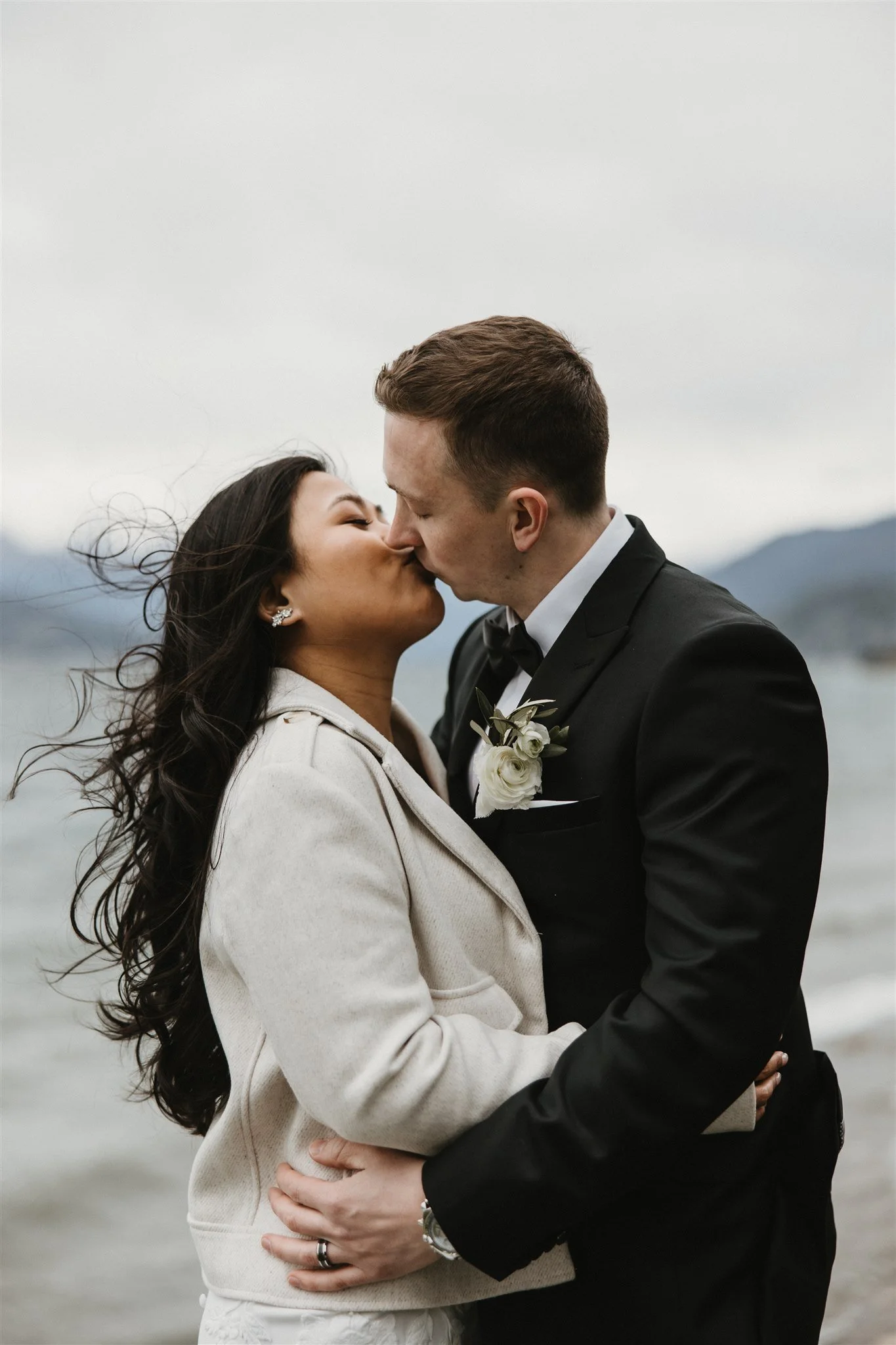 A bride and groom kissing outdoors near a body of water with mountains in the background.