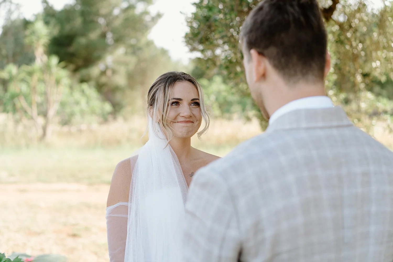 A woman with blonde hair in an updo and a white dress looks at a man with brown hair in a gray suit outdoors during daytime.