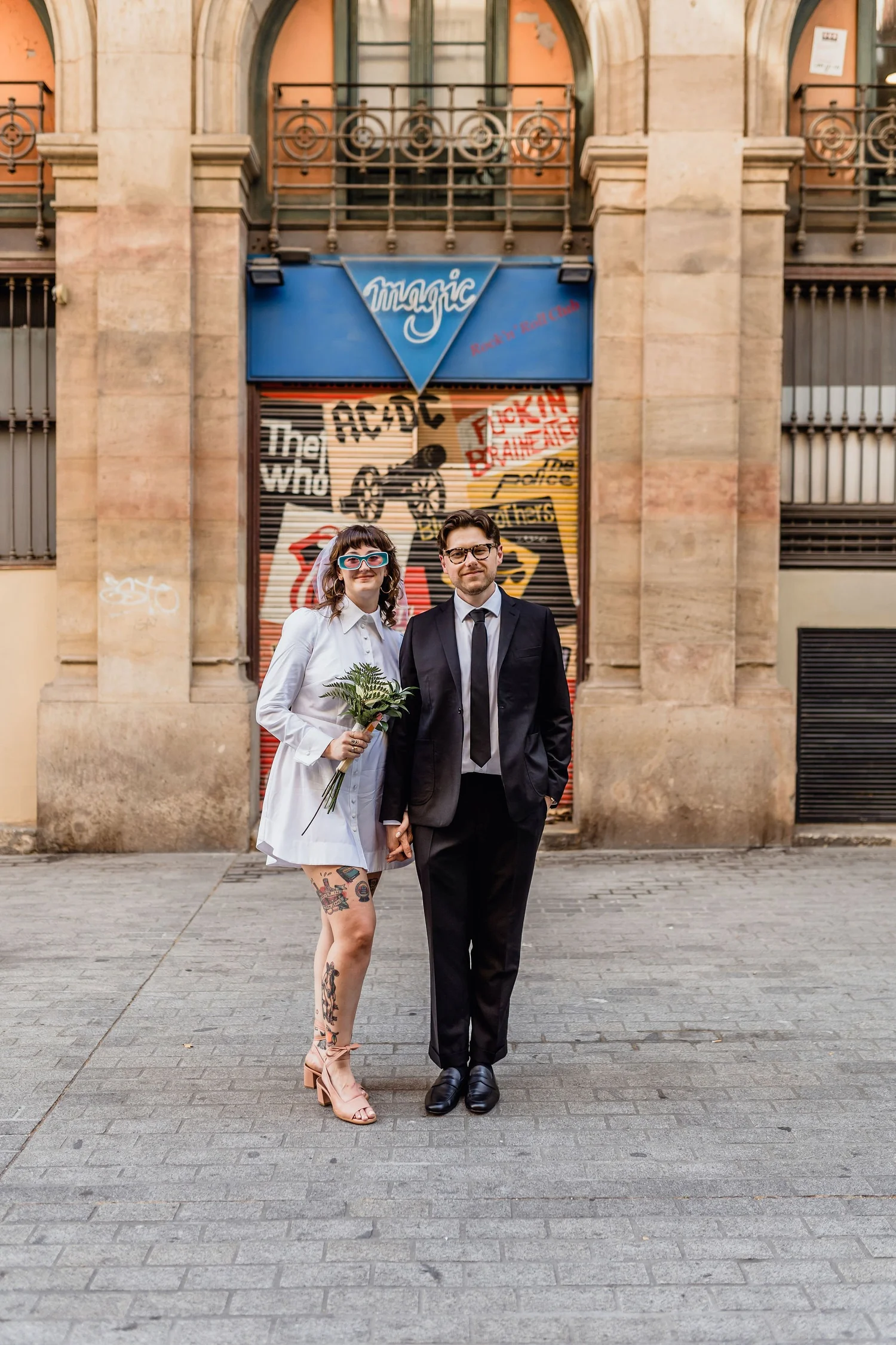 A woman and a man are standing on a cobblestone street in front of a colorful, graffiti-covered building with a blue sign that reads 'Magic.' The woman has tattoos on her legs and is wearing a white shirt dress, pink high heels, and sunglasses, and i