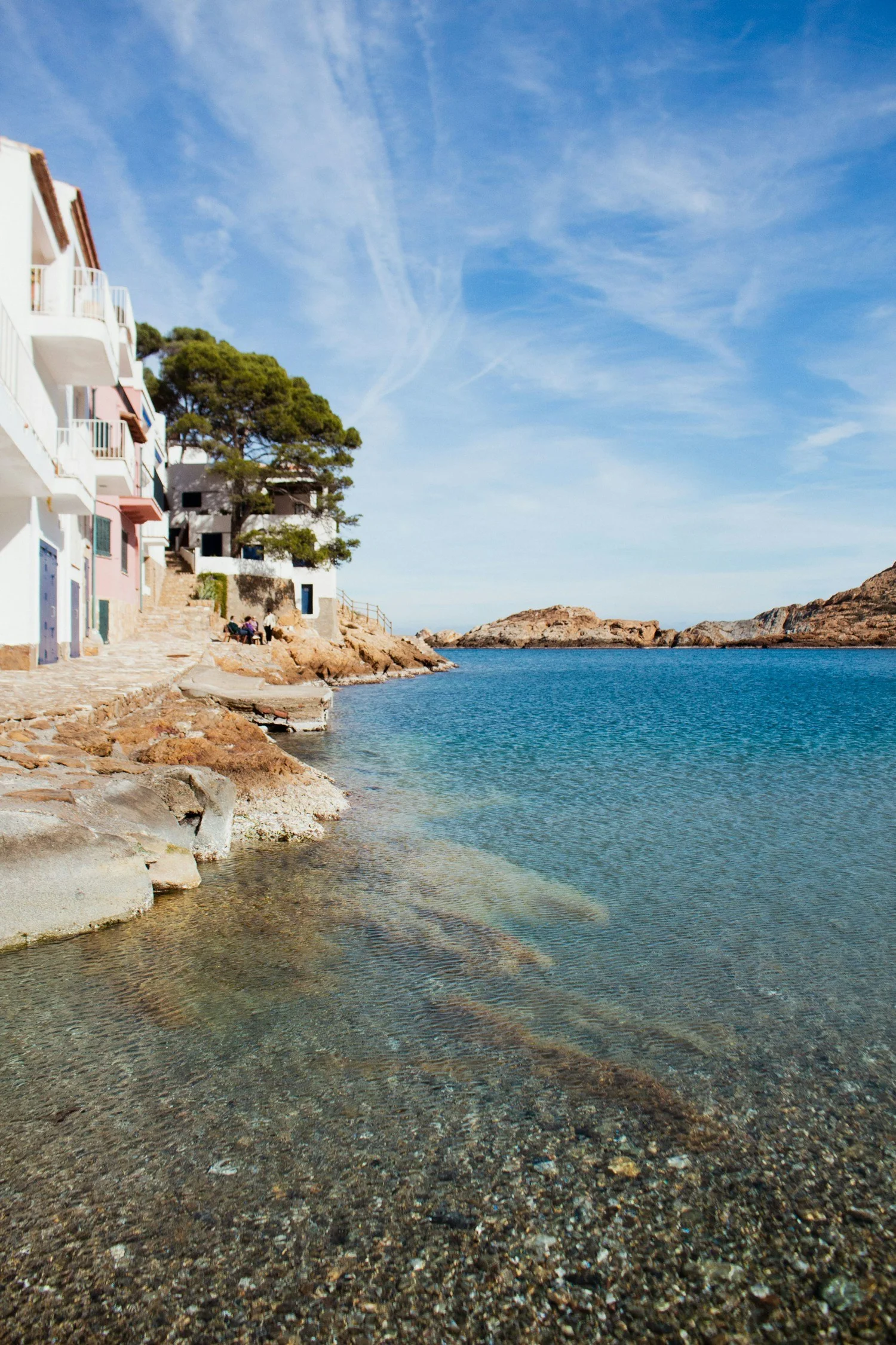 A peaceful coastal scene with clear water, rocky shoreline, colorful buildings on the left, a large tree, and blue sky with wispy clouds.