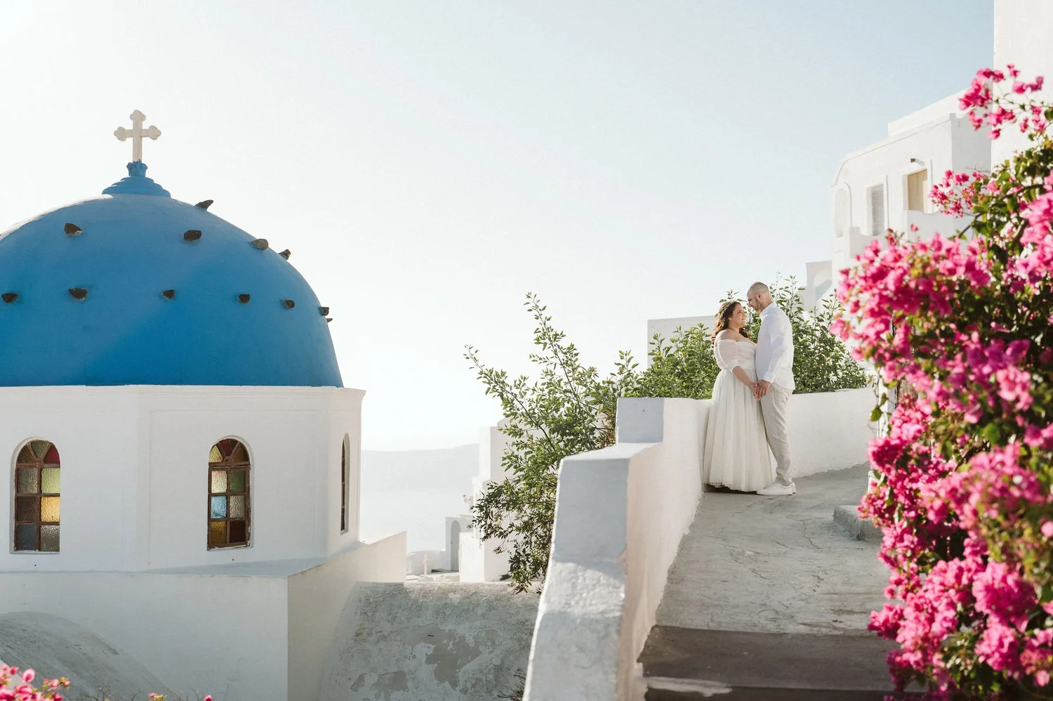 A couple dressed in white wedding attire standing on a white ledge, gazing into each other's eyes, in front of a bright blue-domed church with a cross on top and colorful windows, with pink flowers in the foreground on a sunny day in Greece.