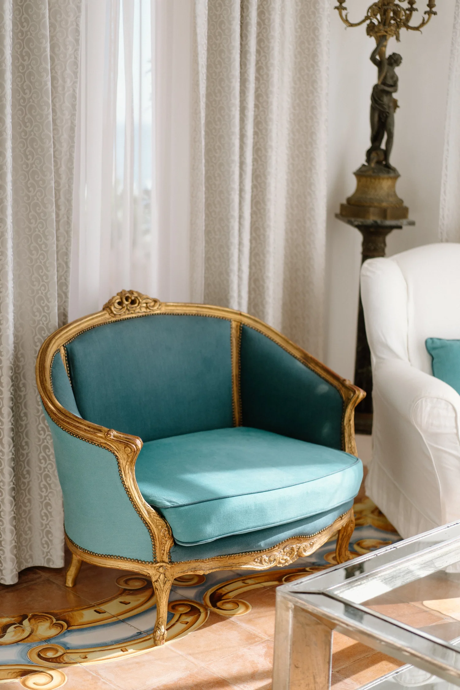 An elegant room with a vintage teal and gold upholstered chair, white curtains, a sculpture on a wooden stand, and a white sofa with a teal pillow.