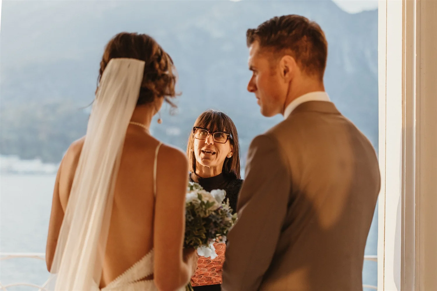A couple getting married, standing close together, facing each other during their wedding ceremony on a balcony with a view of the ocean and mountains in the background. An officiant stands between them, holding a bouquet of flowers.