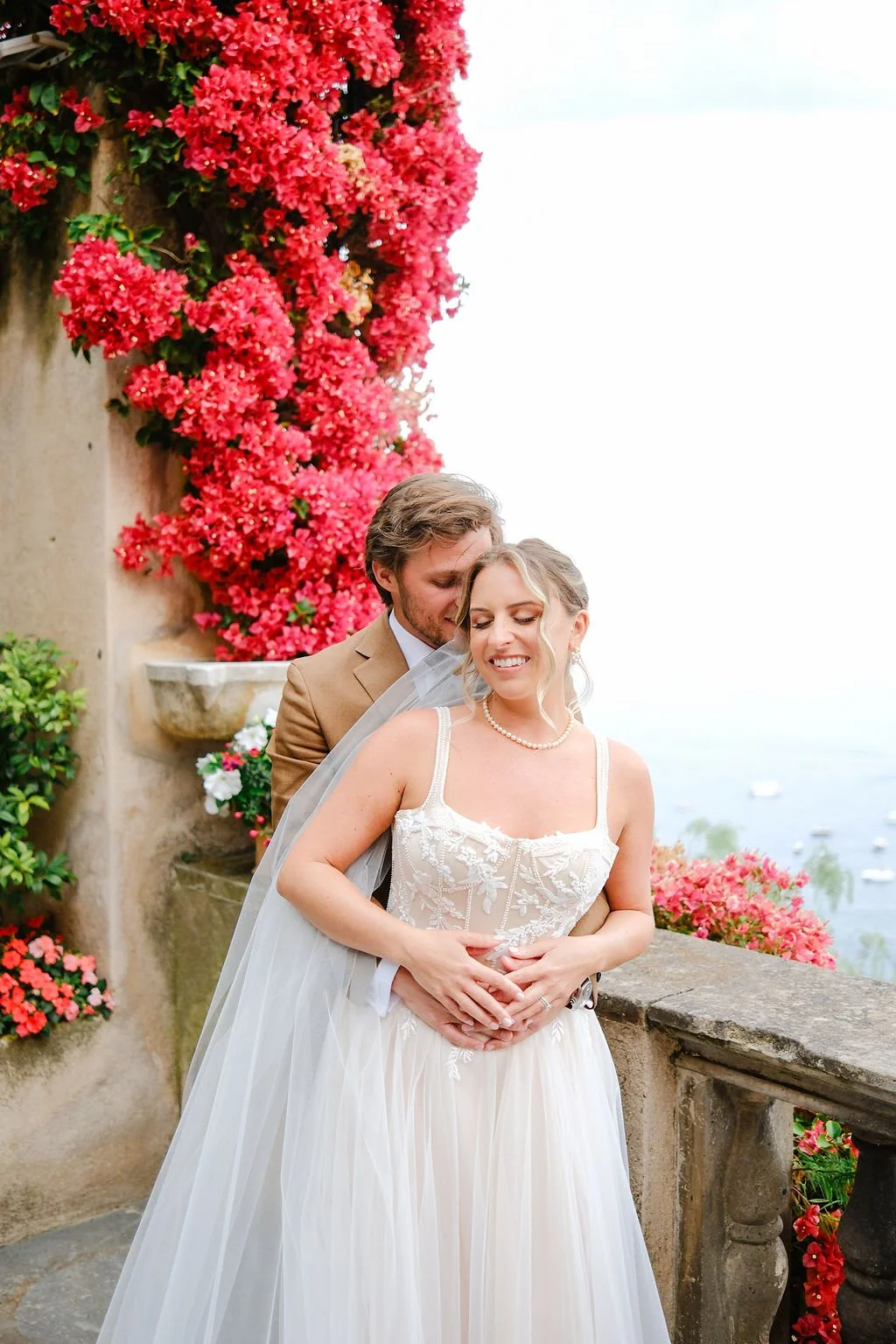 Bride and groom embracing on a balcony overlooking a body of water and boats, surrounded by vibrant pink flowers.