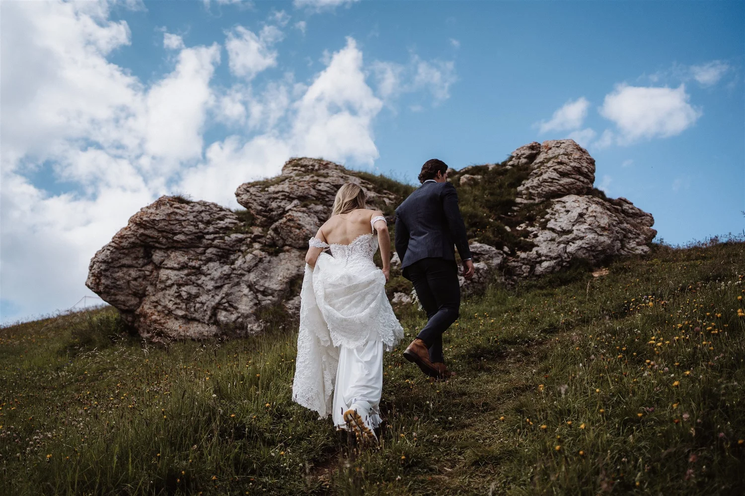 A bride and groom walking up a grassy hill with rocky formations behind them, under a partly cloudy sky.
