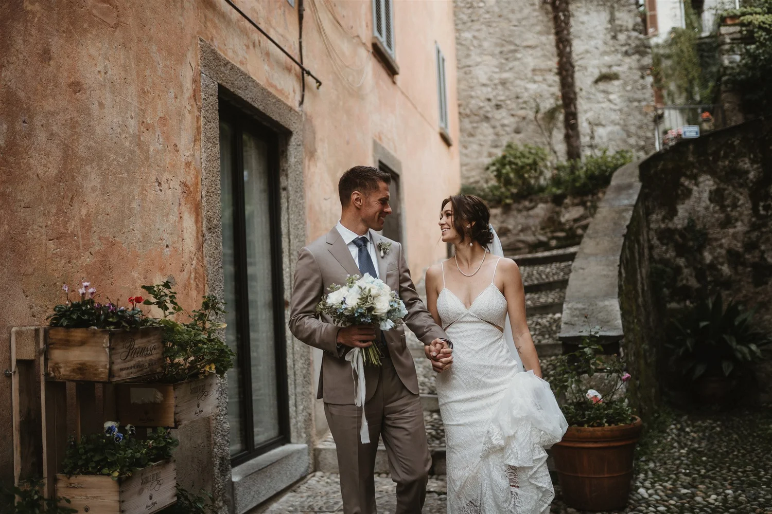 A newlywed couple holding hands and smiling at each other outside on a cobblestone street, with stone buildings and potted plants around them.