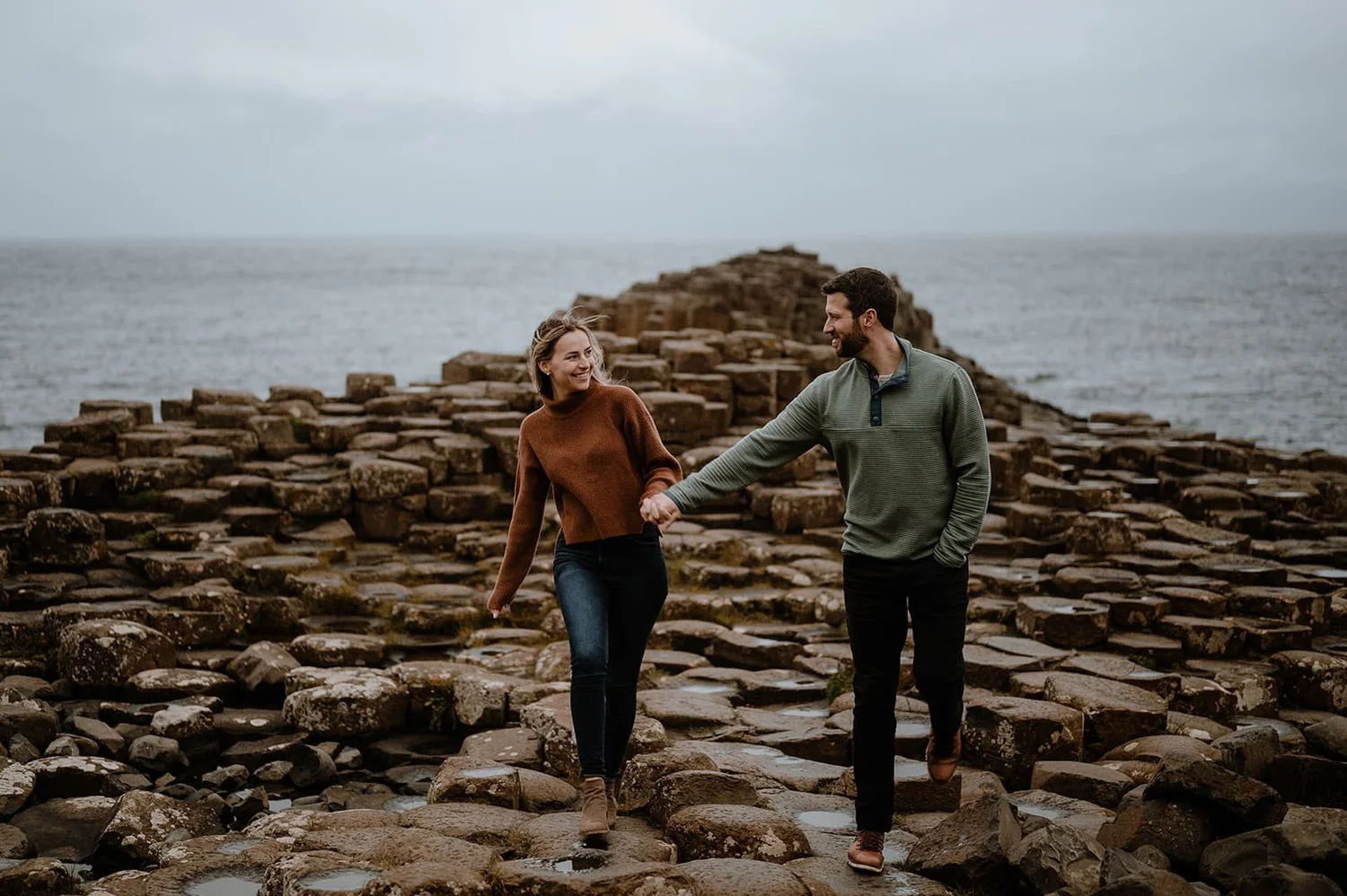 A couple walking hand in hand on rocky path by the ocean during cloudy day.
