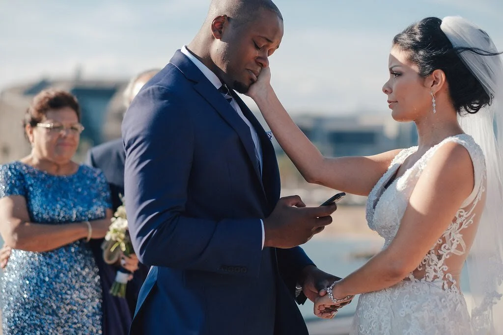A bride and groom holding hands during their wedding ceremony on a sunny day, with a woman in a blue dress and glasses standing in the background.