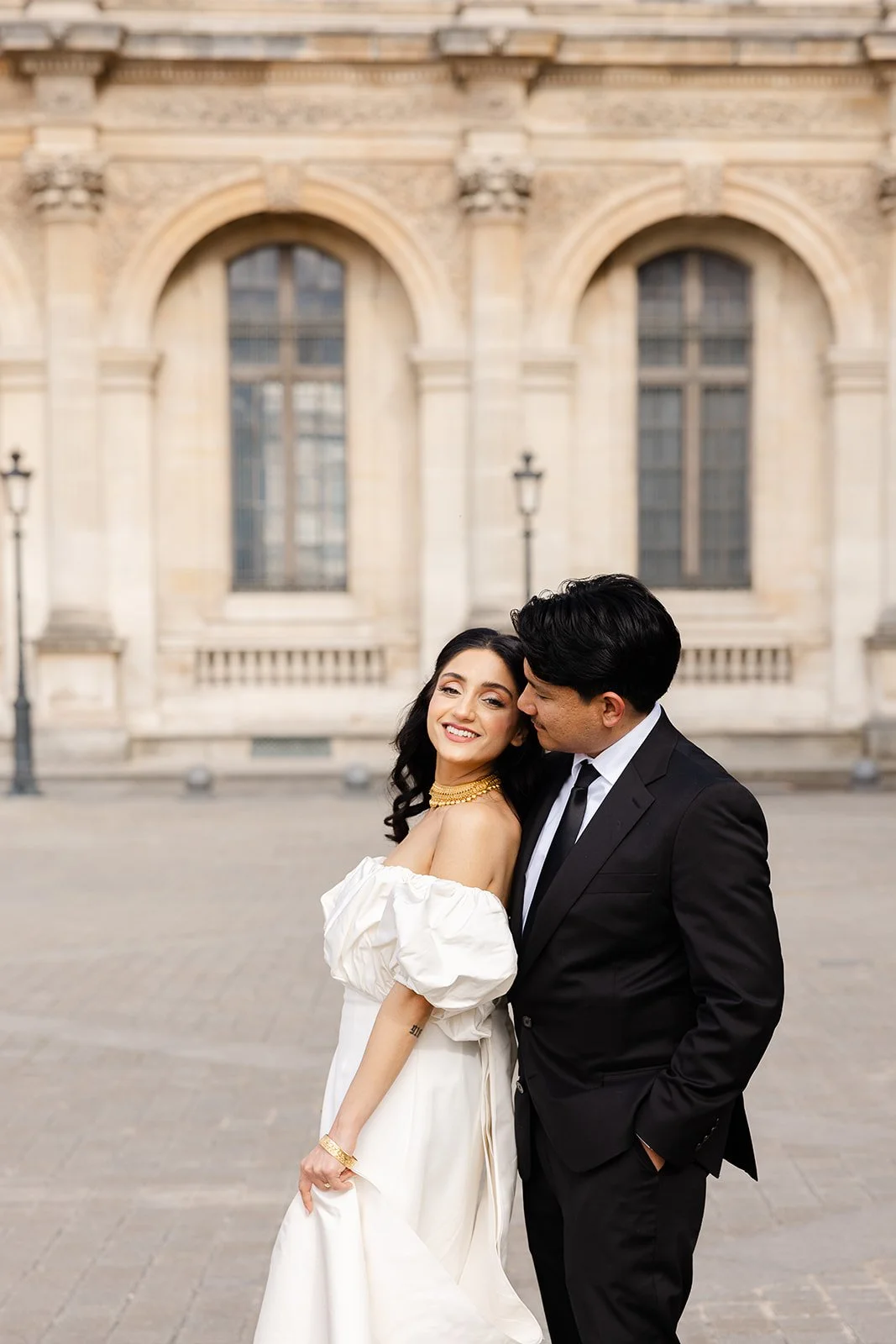 A happy couple dressed in formal attire posing outdoors in front of a historic stone building with large arched windows and street lamps.