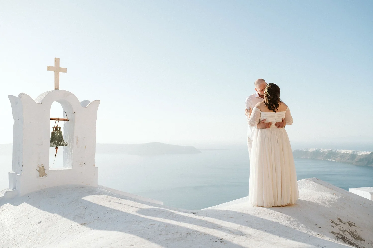 A couple dressed in wedding attire sharing a kiss and embrace on a white terrace in Santorini, Greece with a view of the sea in the background.