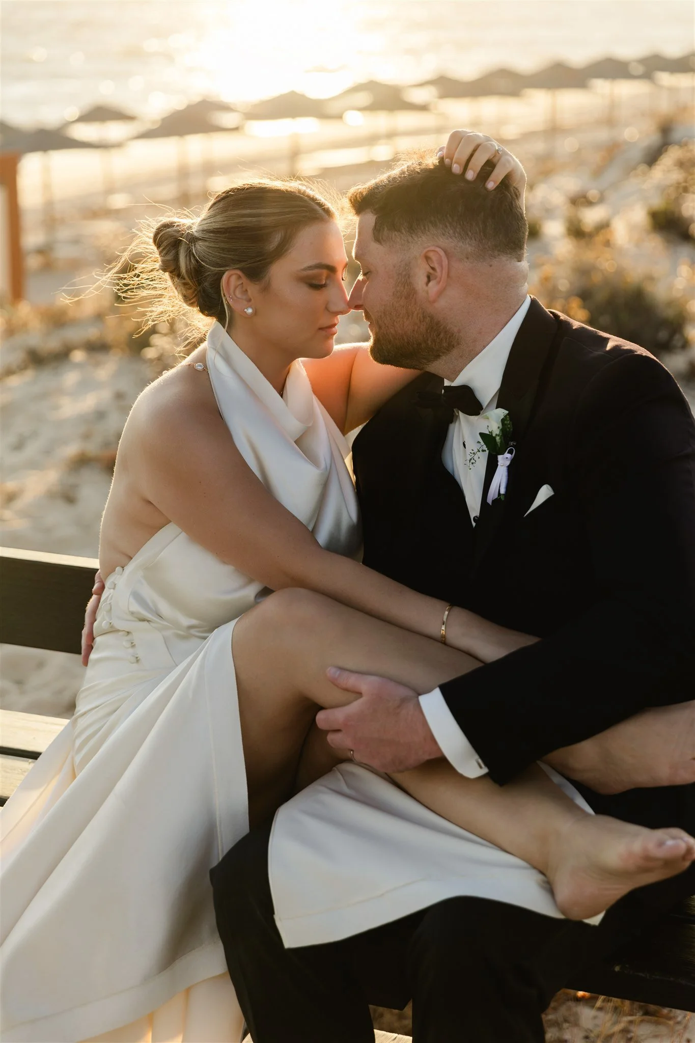 A bride and groom sit close together with their foreheads touching on a beach during sunset, embracing affectionately.