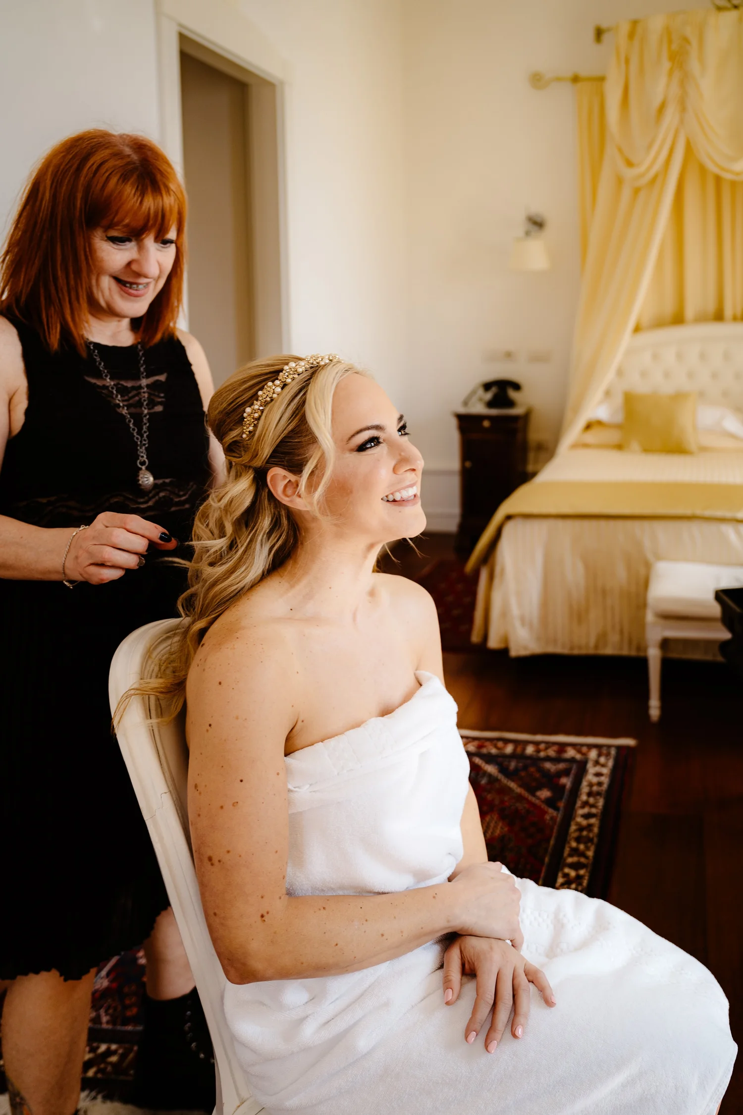 A bride sitting with a towel wrapped around her, smiling as a woman styles her blonde hair, which is adorned with a pearl headband, in a warmly decorated bedroom with yellow and beige tones.