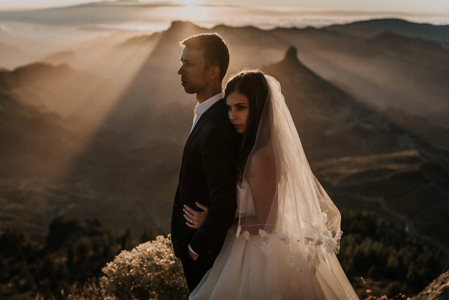 A bride and groom standing close together on a beach at sunset, with the groom facing left and the bride resting her head on his shoulder, wearing a wedding dress and veil.